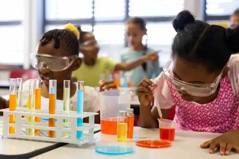 Students wearing safety goggles conducting chemistry experiments with colorful liquids in test tubes at a classroom laboratory station